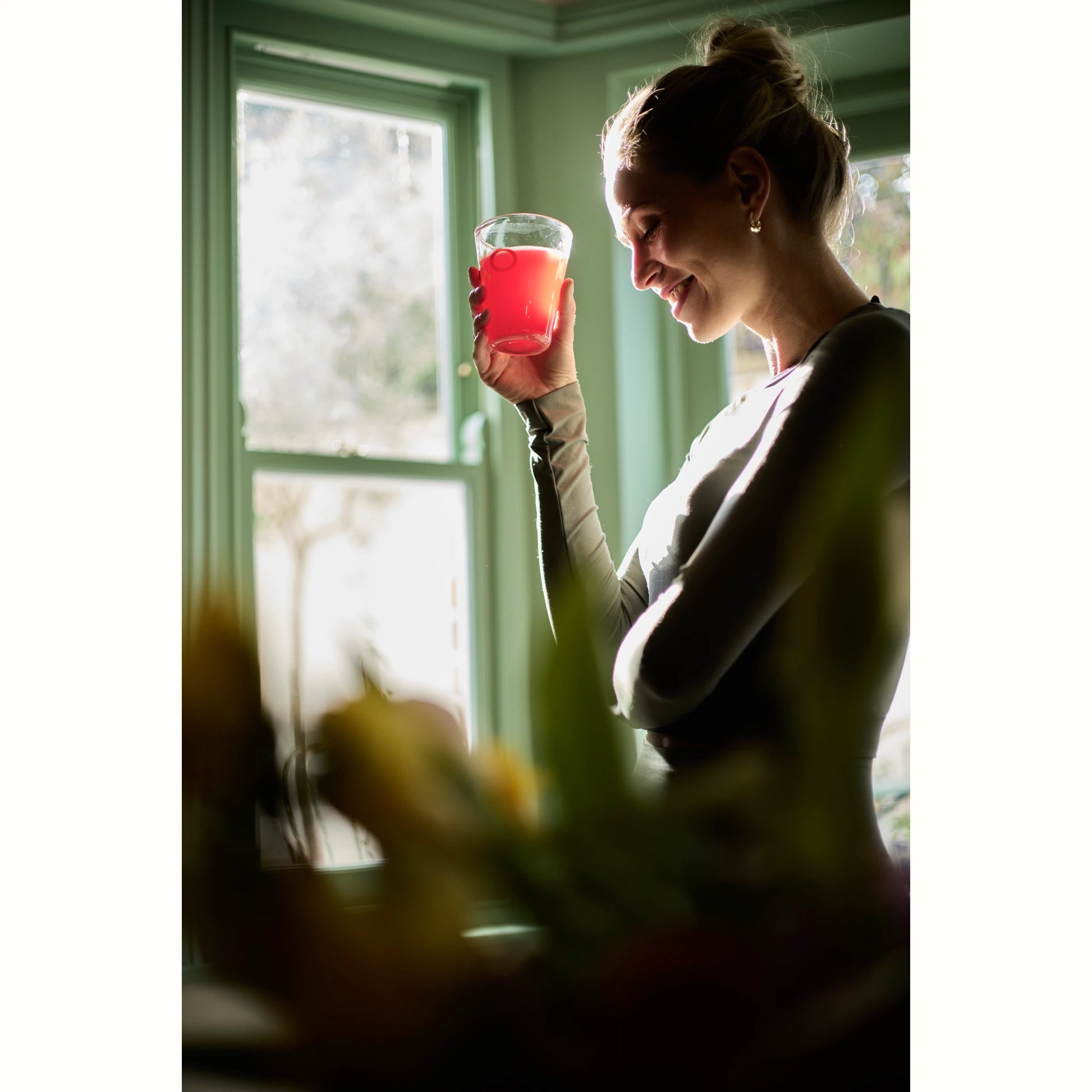 Vrouw glimlacht terwijl ze een glas Aminowise drank vasthoudt bij het raam – natuurlijke ondersteuning bij migraine en hoofdpijnklachten.