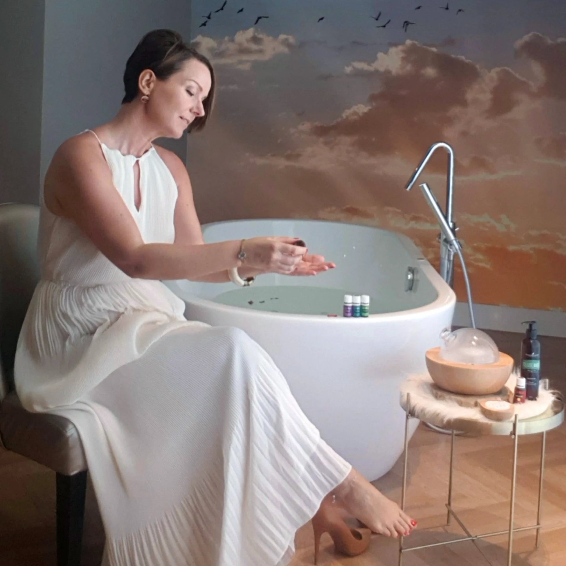 Suzanne zit bij een bad en druppelt essentiële oliën op haar hand, terwijl een Aria diffuser en andere Young Living producten op een tafeltje staan. Ultieme ontspanning met natuurlijke wellness.