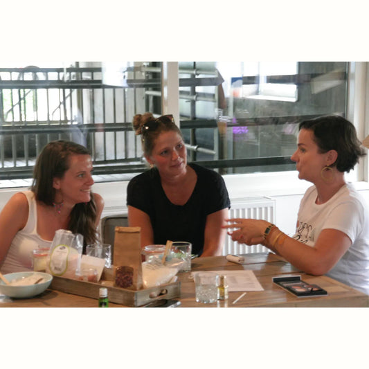 Drie vrouwen in gesprek aan een houten tafel tijdens een holistische coaching bij Suzanne Shares, omringd door natuurlijke producten en essentiële oliën – gericht op het reguleren van het zenuwstelsel voor herstel en balans bij vrouwen boven de 40.