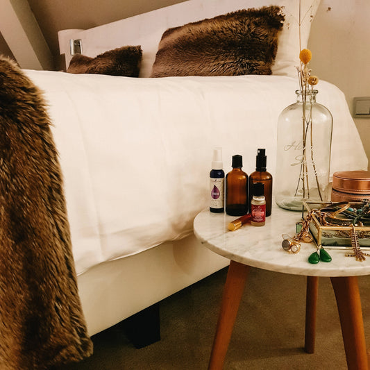 Nachtkastje met natuurlijke slaapondersteuning zoals etherische oliën, sprays en rollers naast een opgemaakt bed – rustgevende slaapkameromgeving voor vrouwen 40+ met slaapproblemen.