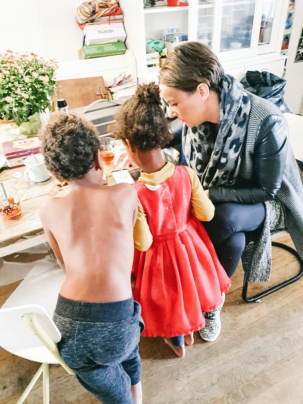 Vrouw begeleidt twee jonge kinderen aan een keukentafel terwijl ze samen naar een telefoon kijken. De sfeer is warm en betrokken. Er staan glazen, bloemen en oliën op tafel.