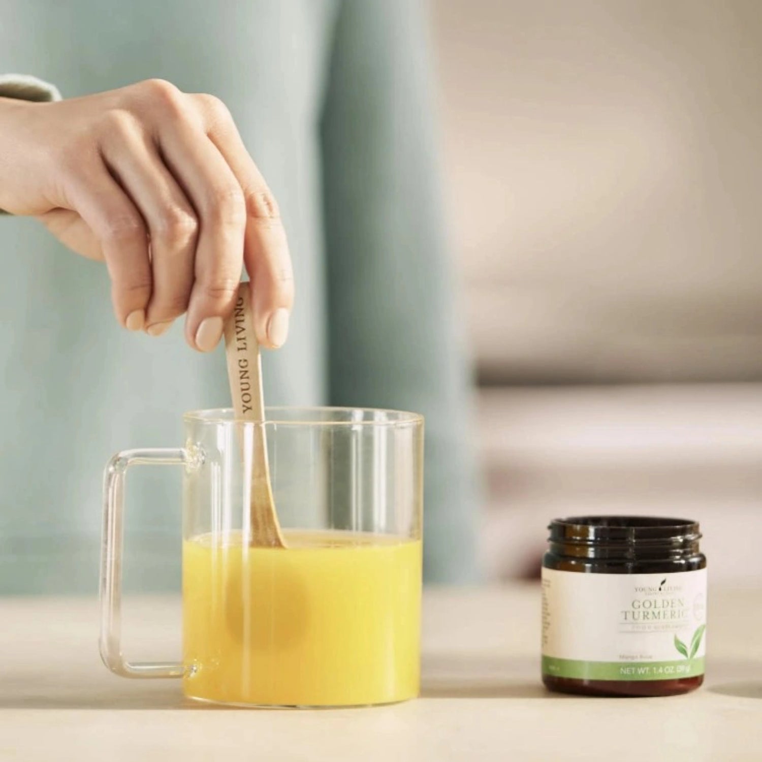 Hand roert Golden Turmeric kurkuma-drank in een glazen mok naast de geopende Young Living-verpakking.