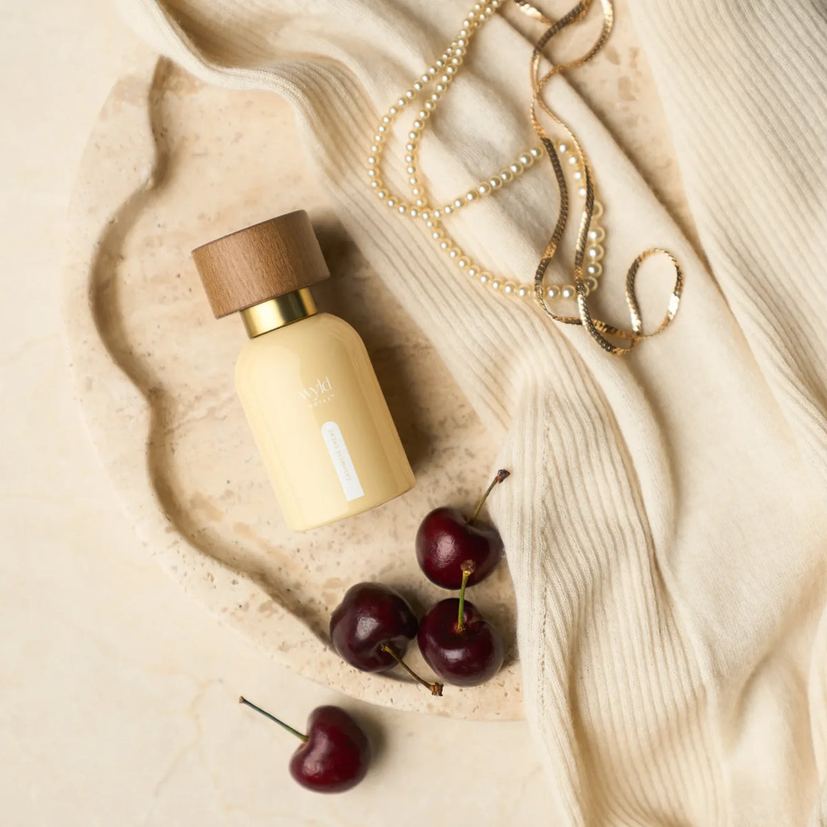 Wyld Notes Cashmere Crème parfum tussen zachte crèmekleurige stof, kersen en sieraden in een elegante lifestyle-setting.