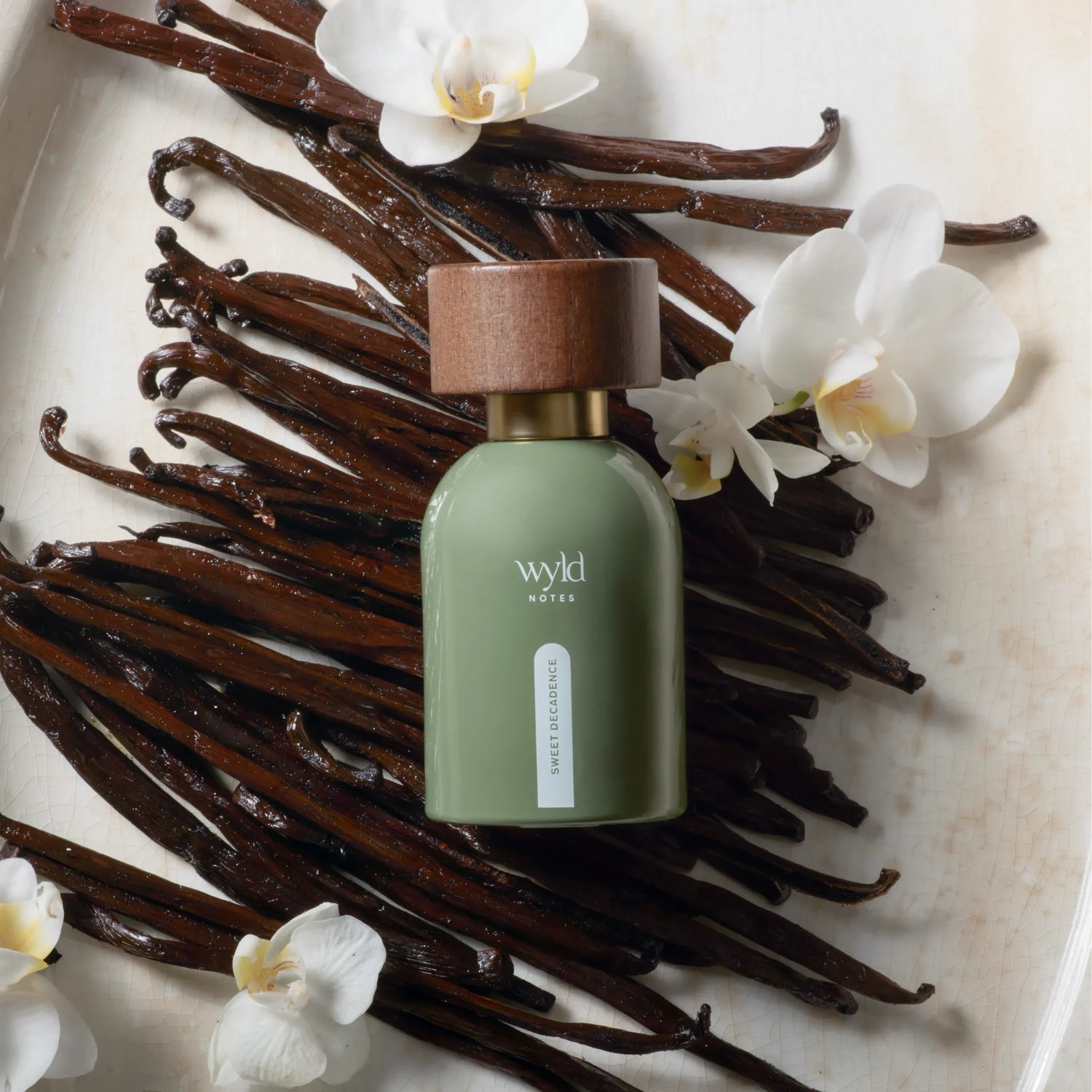Wyld Notes Sweet Decadence eau de parfum in groene flacon, omringd door vanillestokjes en witte orchideeën.